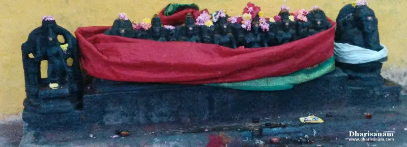 A row of small dark idols with flowers, partly covered by a red cloth, against a yellow wall