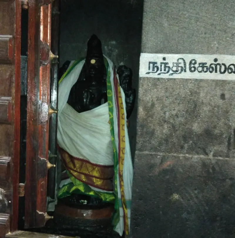 Dark standing figure in white cloth beside a pillar with a Tamil label