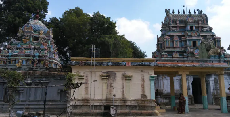 A painted tower at right and a domed shrine at left, with a plain building between; a woman in a sari stands near.