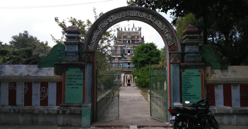 A stone arch with Tamil lettering over an iron gate; a path leads to a coloured tower, a motorbike at right.
