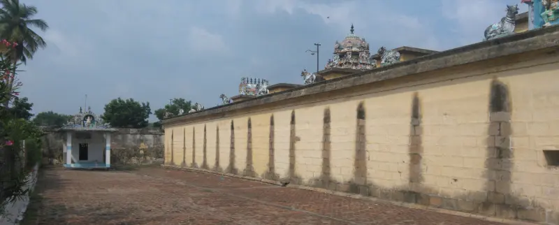 A long brick-paved corridor beside a high compound wall with a small shrine at the far end