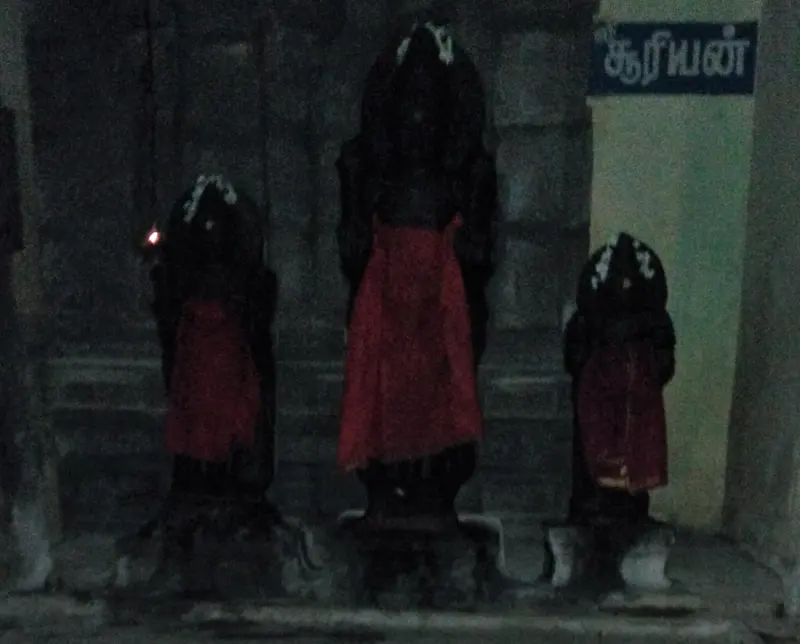 Three dark idols with red cloth stand together in a corridor, with a Suryan board at the right