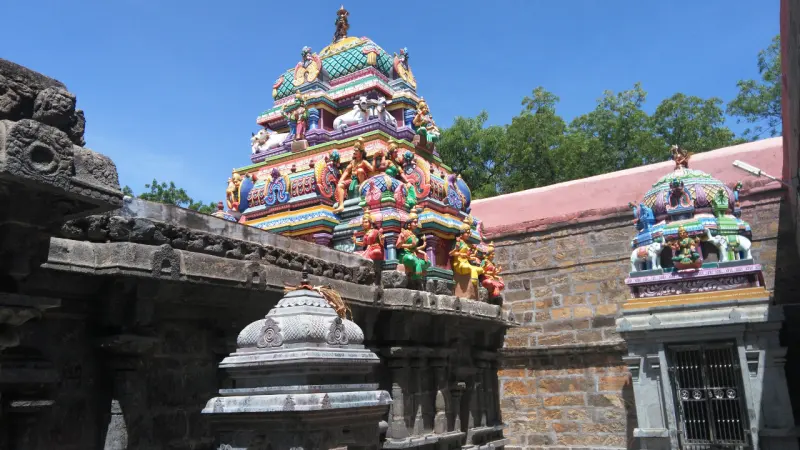 A colourful tower rises behind a carved stone wall, with a small shrine at right and a stone finial in front.