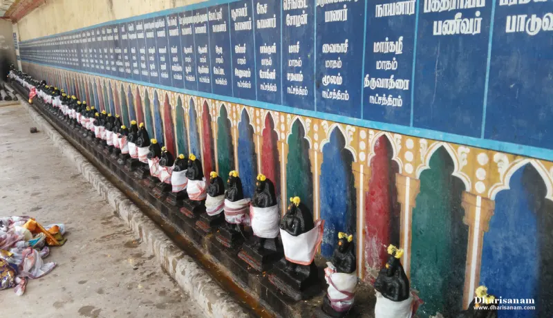 Many small black idols in white cloths on a ledge, with blue Tamil name panels above