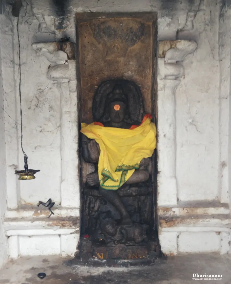 Dark idol seated in a tall niche with a yellow cloth over its lap, and a hanging lamp at the left