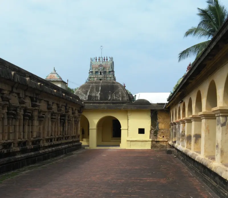 A brick-paved corridor between a carved stone wall and an arched cloister leads to a yellow domed hall.