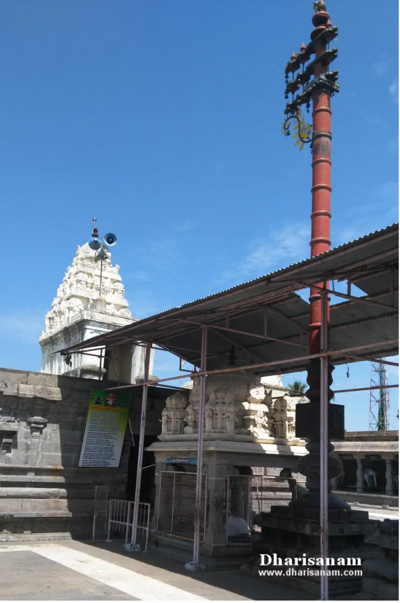 Tall red flagstaff beside a small carved shrine under a metal roof, with a white tower behind