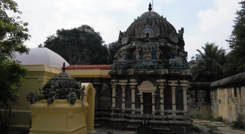A dark stone shrine with a carved dome and a small yellow shrine at left, with trees and a white-domed roof behind.