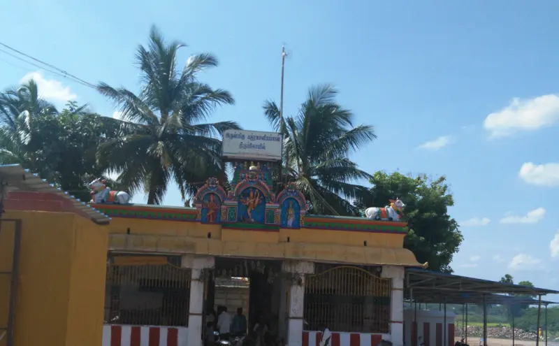 A small shrine entrance with painted figures and a name board, between coconut palms under a blue sky