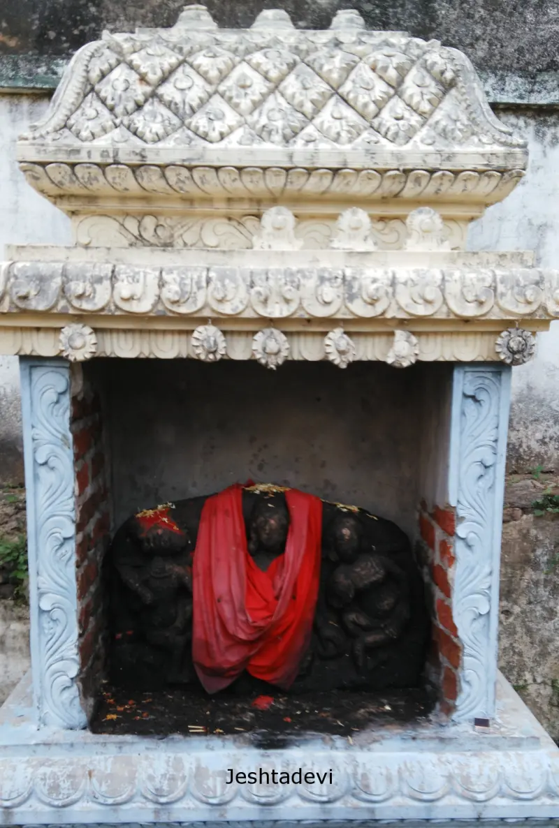 A dark stone carving in a small shrine, a red cloth draped over it; a caption names Jeshtadevi.