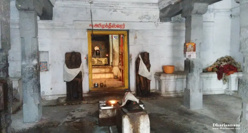 Lit shrine doorway on a white wall flanked by two draped dark stone figures, with a small lingam in the foreground