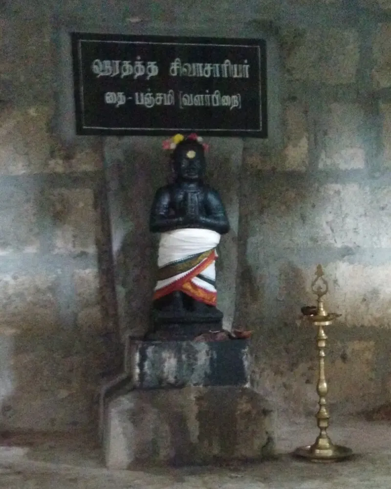 A black idol with folded hands stands beside a lamp, under a Tamil name board on the wall