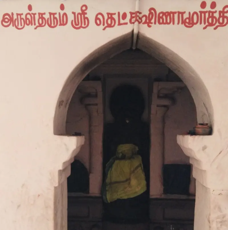 A deity draped in yellow cloth stands in an arched niche, with Tamil lettering above naming Dakshinamurthy.