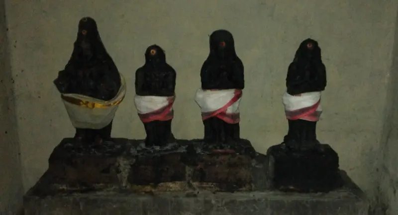 Four small dark stone idols with red dots on their foreheads, wrapped in white cloths, standing on a stone platform.