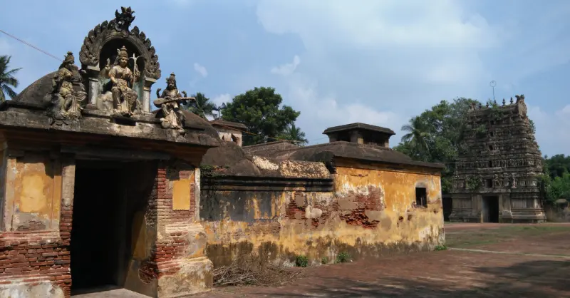 An old gateway with stone figures on top beside a yellow-stained wall; a stone tower stands among trees.