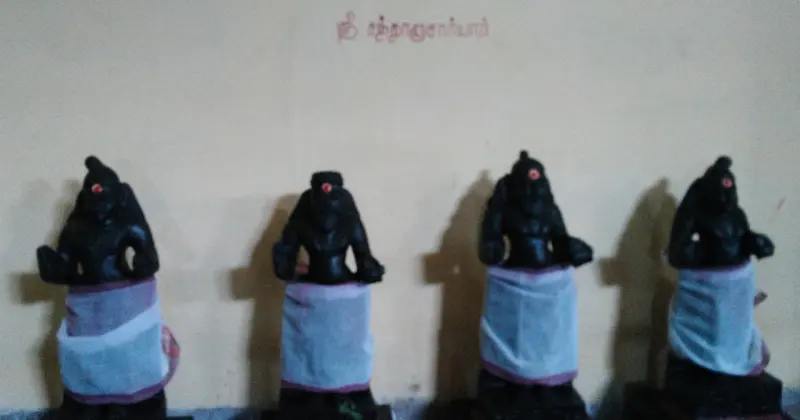 Four dark idols in white cloth standing in a row before a yellow wall with a painted name above.