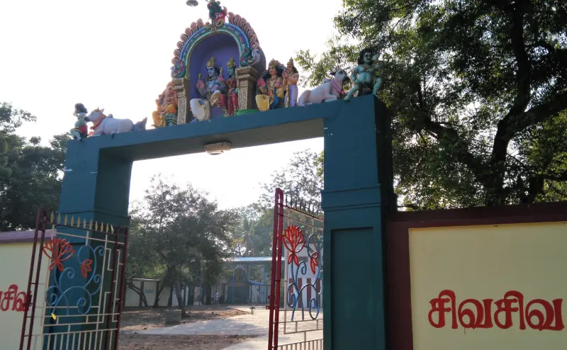 A green arch over a gate, topped with painted figures of deities and bulls, with trees behind.