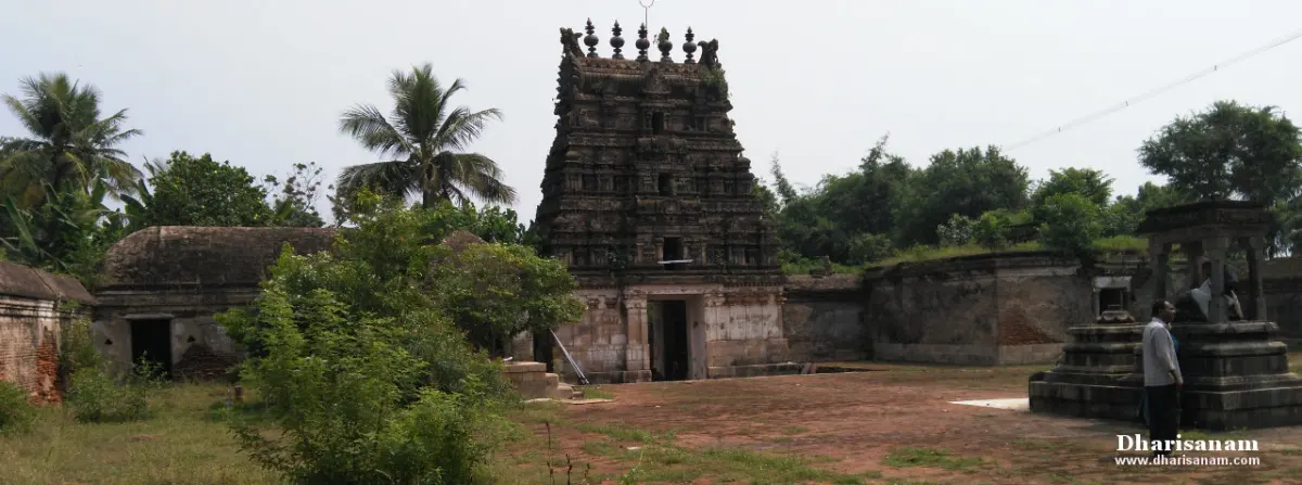 A weathered stone gopuram rises over a brick-paved courtyard, with palm trees behind and a stone pavilion at right.