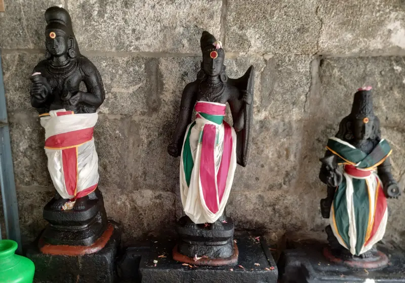 Three dark stone idols in a row against a stone wall, each dressed in white, red and green cloth.