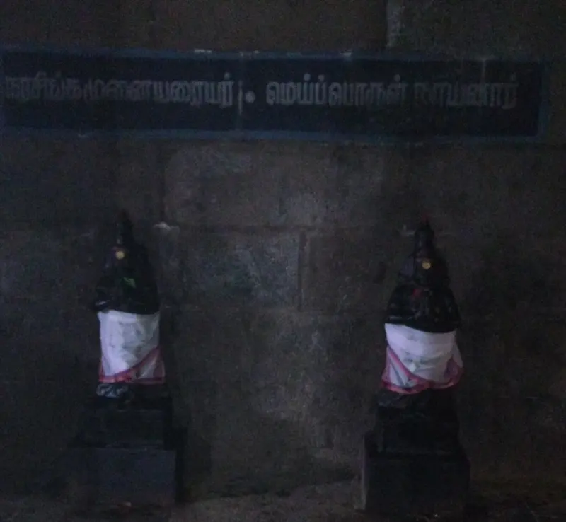 Two small dark idols in white cloth stand against a stone wall under a board with Tamil names