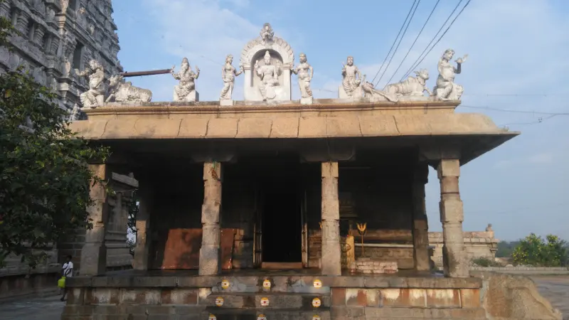 An open stone mandapam with square pillars and white figures standing along its roof edge