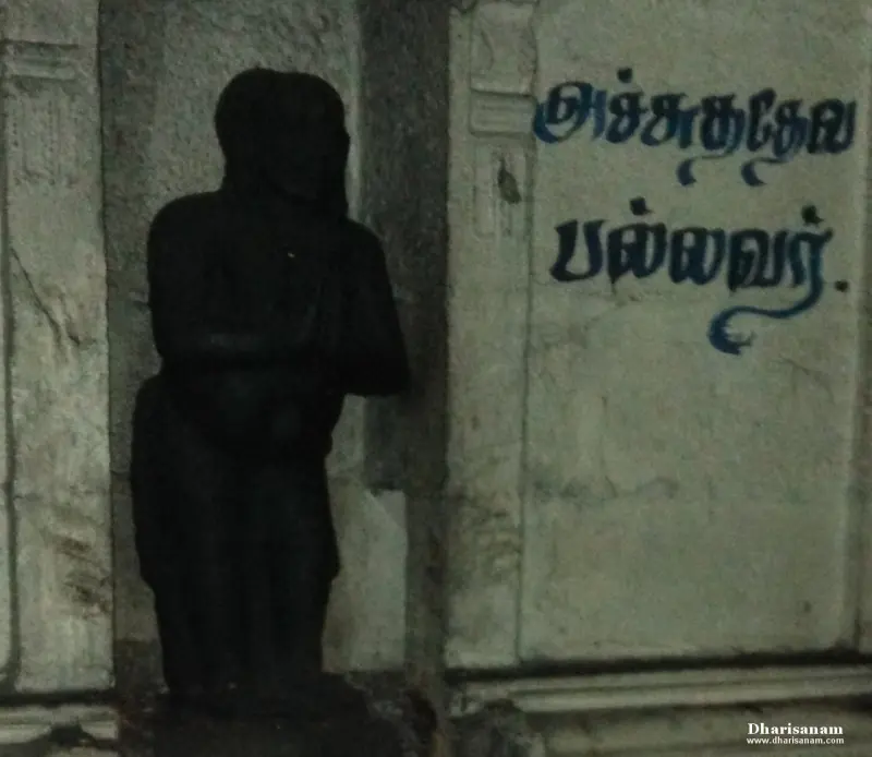 A dark stone figure standing beside a wall on which his name is painted in blue Tamil letters
