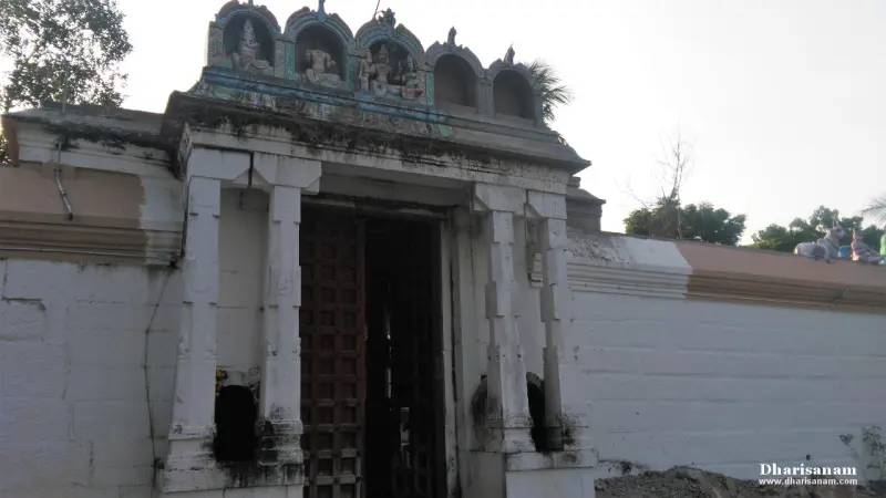 A white pillared arch gateway with a wooden door and small painted figures on top, set in a plain white wall
