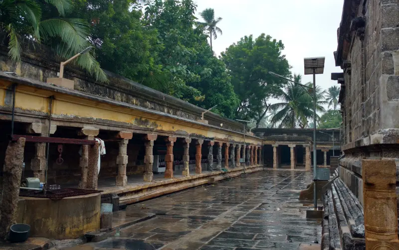 A long open corridor with stone pillars beside a wet paved courtyard, under trees.