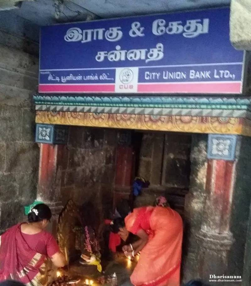 Devotees bending to light lamps at a small stone shrine under a blue name board