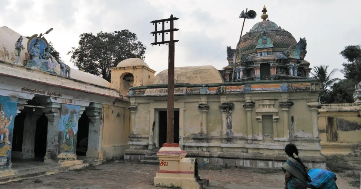 A tall flagstaff in a courtyard before a pillared hall and a domed sanctum tower; a woman stands at lower right.