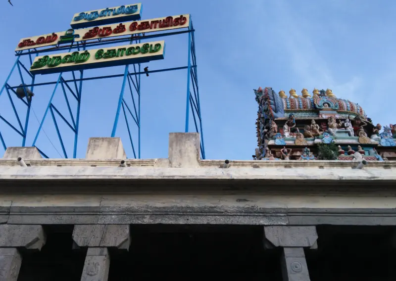 A blue frame with a Tamil name board beside a colourful sculpted tower top, above a stone wall