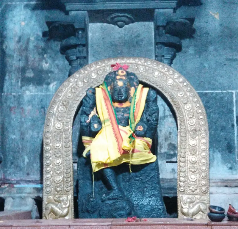 A dark stone idol in yellow cloth beneath an arch of gold-coloured metal, with small lamps at the base.