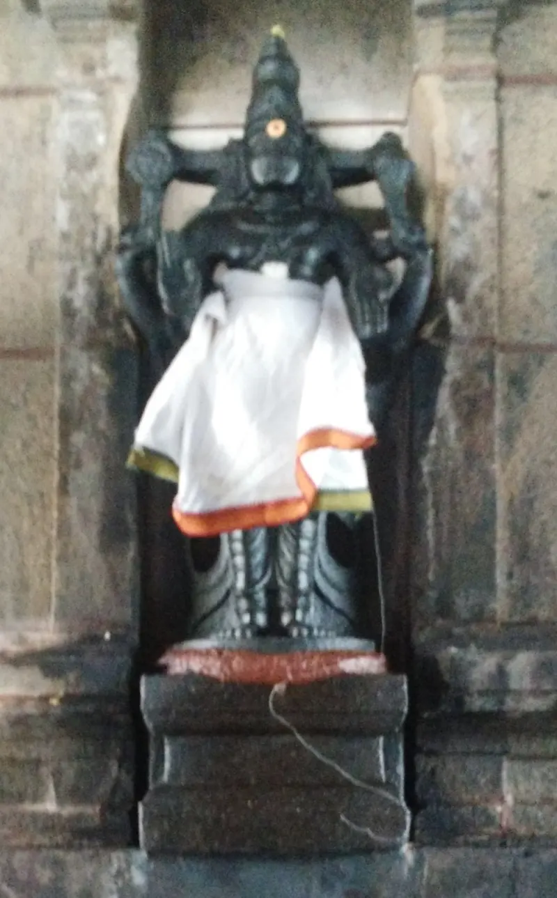 A black stone figure with raised arms stands in a niche in a stone wall, wearing a white cloth with an orange border.