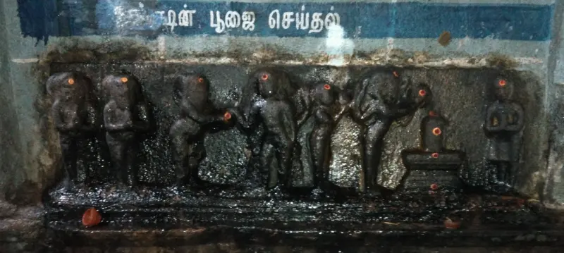 A row of dark carved figures in relief, with a Lingam at the right end and a Tamil caption on the wall above.