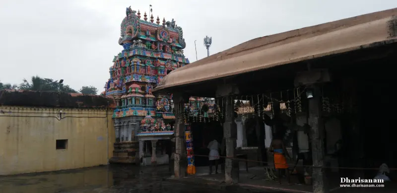 A tiered gopuram seen from the courtyard, with a tiled hall roof at right and people in the shade