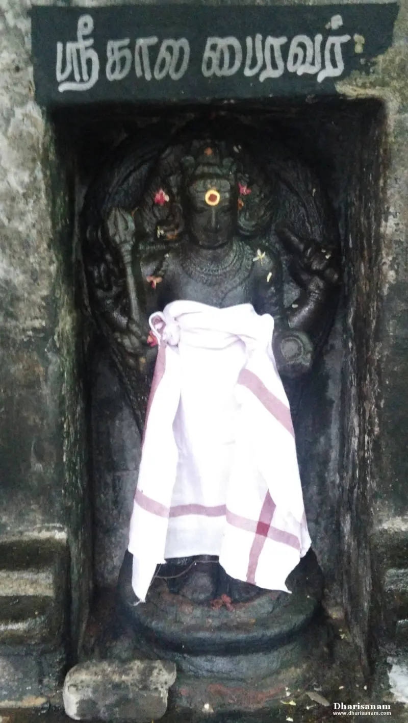 Dark stone Kala Bairavar holding weapons in a niche under a name board, wearing a white cloth