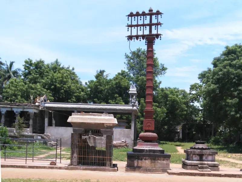 A tall brown flagstaff with bells stands beside a small barred shrine, with trees and a pillared hall behind.