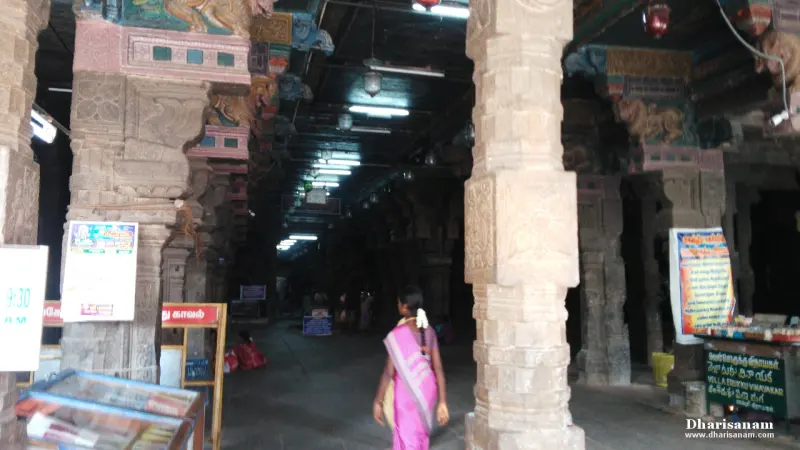 A dim stone hall with carved pillars, a woman in a pink sari walking through it