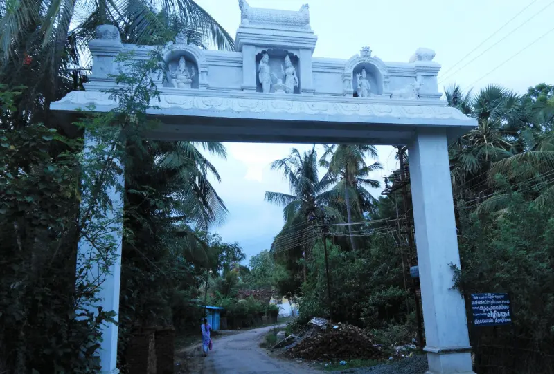 A white arch with sculpted figures in niches spans a narrow road among coconut palms, with a signboard at lower right.