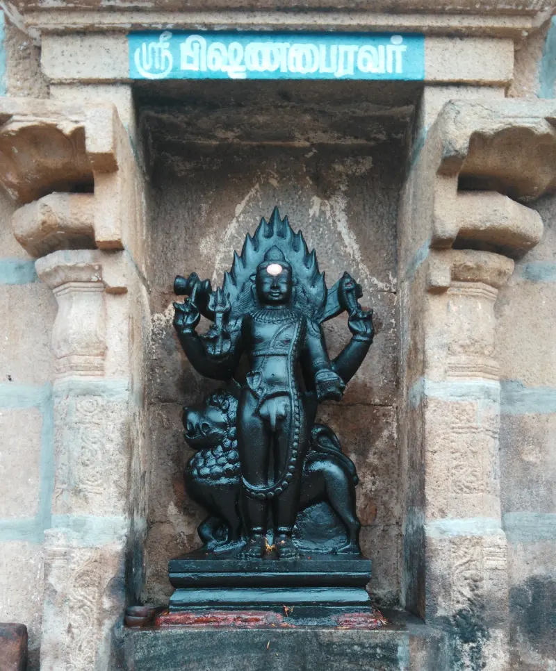 Dark figure with a flame-like crown on a low base, under a blue Tamil name board