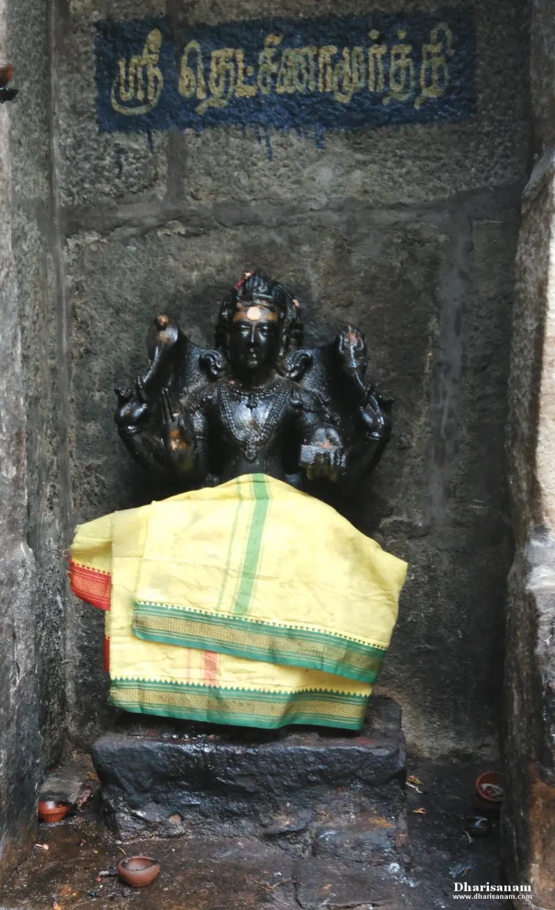 A dark four-armed idol seated on a stone base in a niche, with a yellow cloth over the lap and small oil lamps below