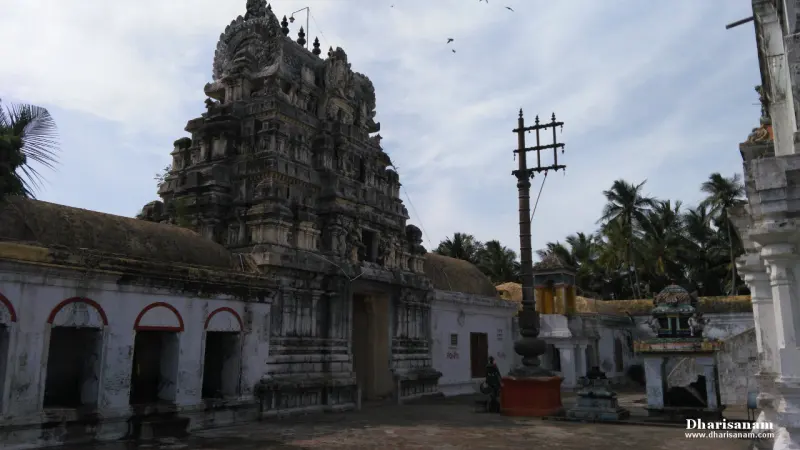 A dark stone gopuram beside a tall flagstaff in an open stone-paved courtyard with palm trees behind