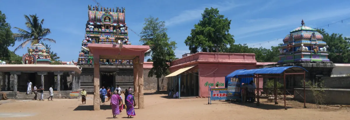 A sandy courtyard with devotees, a stone porch, and a painted tower and a smaller domed shrine behind.