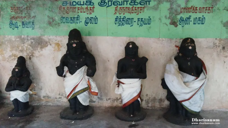 Four black idols in white cloth stand against a wall under a green board naming the saints and their months