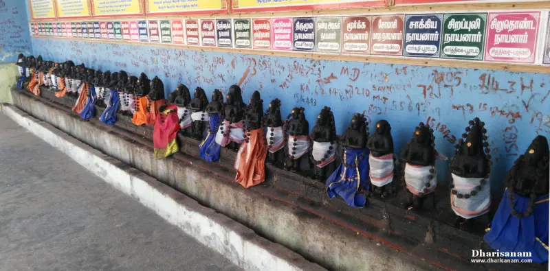 Many small dark idols in cloth wraps on a long ledge, each with a coloured name board on the wall above.