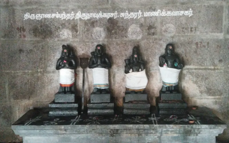 Four small black idols in white cloth on a stone platform, with Tamil names painted on the wall