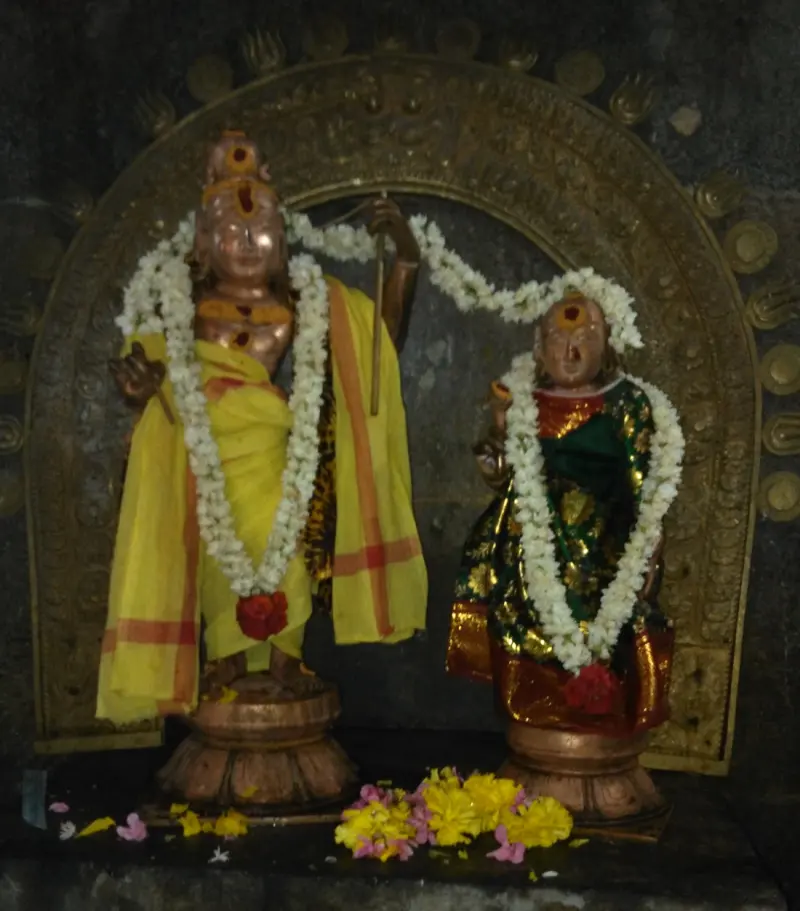 Two bronze idols, a god holding a staff and a goddess, both garlanded with jasmine, beneath a bronze arch.