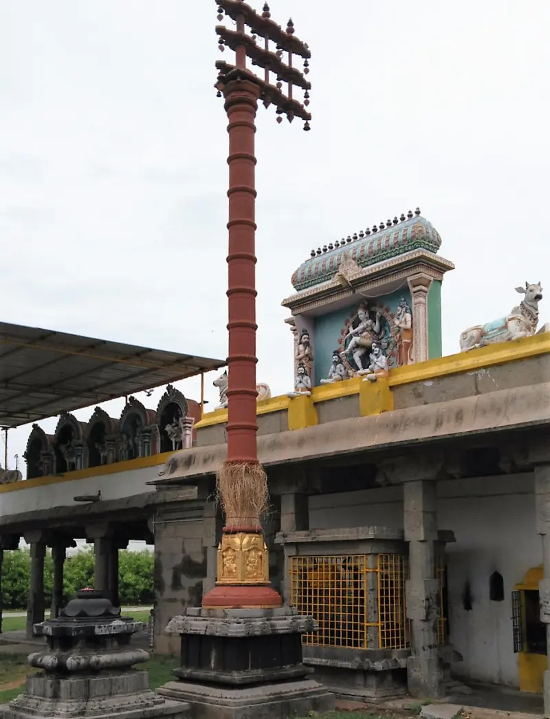 A tall red flag post with a gold base beside a hall whose roof shows a dancing figure and a Nandi.
