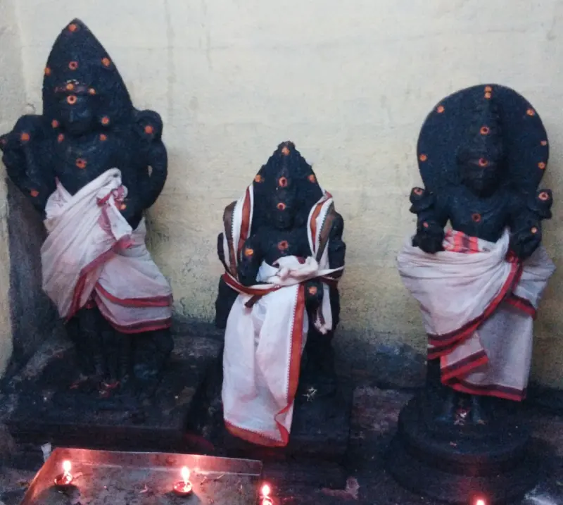 Three dark stone idols in white veshtis with oil lamps lit before them