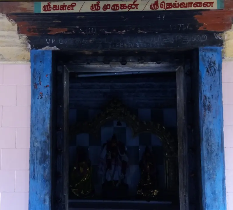 Dim shrine under a Tamil board naming Murugan with Valli and Deivanai, with three images inside
